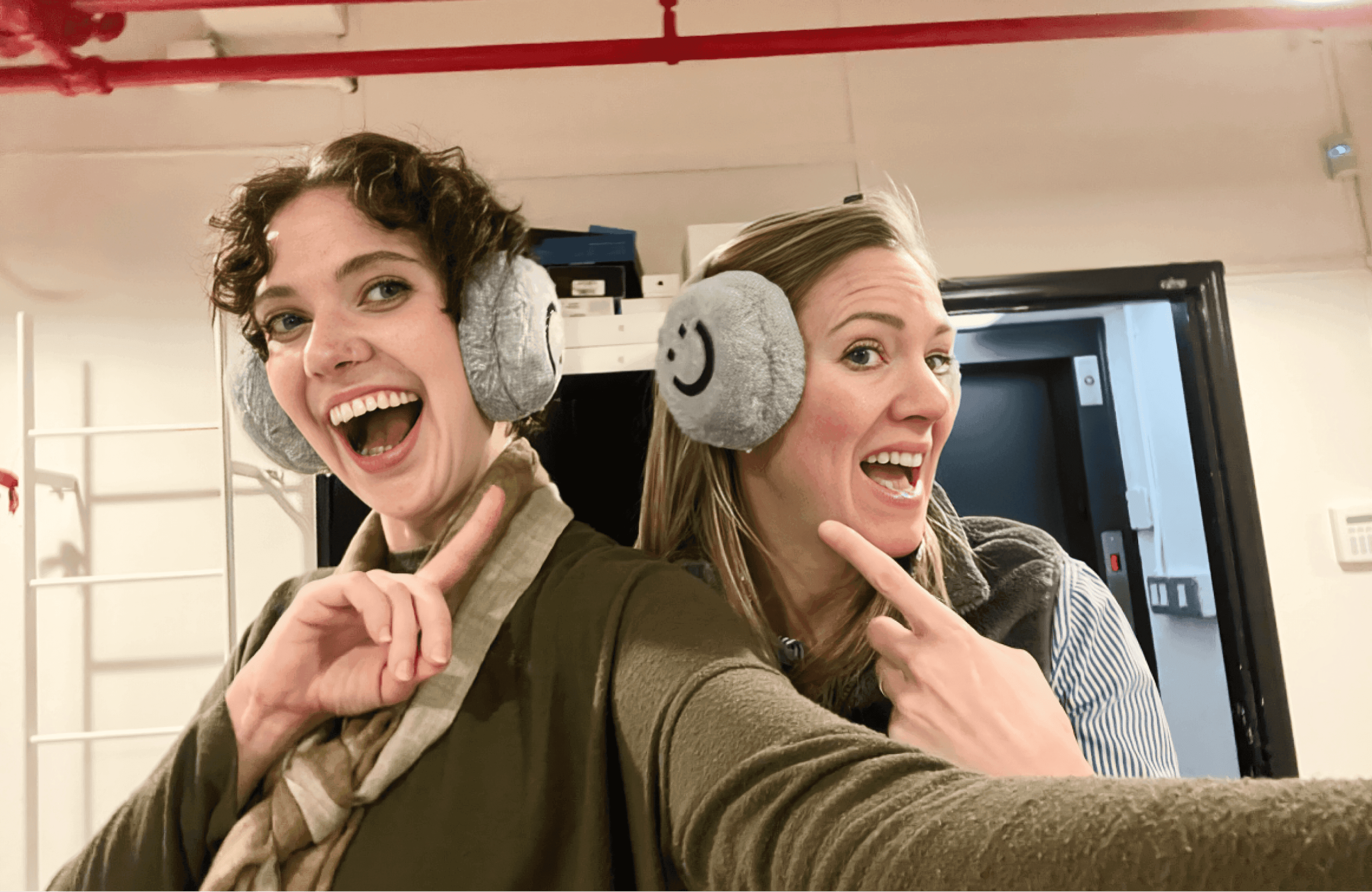 Selfie of two people, with large smiles, each pointing at fuzzy earmuffs they're wearing that feature the Dandi smiley logo.
